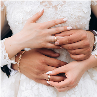 Vor dem Oberkörper der Ehefrau im Brautkleid hält das Ehepaar sich gegenseitig die Hände an den Eheringen fest